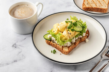 Scrambled eggs on grilled toast with salad leaves, cheese and micro greens on plate on white marble background. Delicious breakfast, brunch, snack