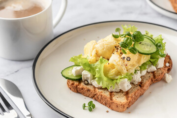 Scrambled eggs on grilled toast with salad leaves, cheese and micro greens on plate on white marble background close up. Delicious healthy breakfast