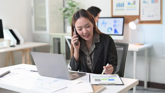 A woman is talking on her cell phone while sitting at a desk with a laptop and a stack of papers