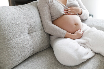 Pregnant woman in cozy clothes hugging baby bump and relaxing on grey couch in modern living room. Fertility and maternity. Happy Mothers day. Love and care concept