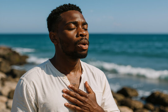 Young man breathing deeply with closed eyes, practicing mindfulness on a rocky beach with the sea in the background