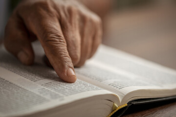 Naklejka premium Close-up, elderly man reading book following text with finger. Senior male reading book at home.