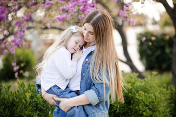 Fototapeta premium Young beautiful mother holding smiling baby girl 3-4 year old posing over flowers in park outdoors. Motherhood. Happiness.