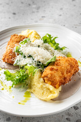A gourmet dish featuring crispy breaded chicken, mashed potatoes, fresh greens, and grated cheese, garnished with a drizzle of green sauce, presented on a white plate with a textured grey background.