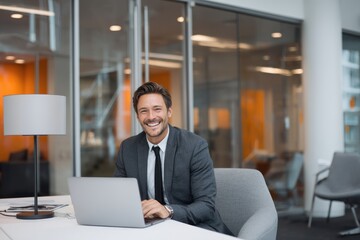Smiling man in suit sits at desk with laptop in office environment.