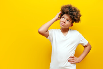 African American puzzled teenage boy in white t-shirt thinking and doubting on yellow isolated background, thoughtful insecure curly schoolboy kid scratching head and looking at copy space