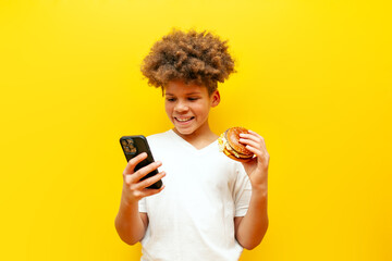 cheerful curly african american boy holding burger and using smartphone online on yellow isolated background, hungry teenager ordering fast food by phone