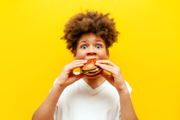 surprised curly african american boy eating burger on yellow isolated background, shocked hungry teenager holding cheeseburger and advertising fast food in amazement