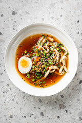 A bowl of ramen with noodles, boiled egg, green onions, carrots, and sesame seeds in a rich broth, presented on a textured gray surface. Warm, appetizing, and authentic Asian cuisine.