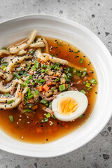A bowl of ramen with noodles, boiled egg, green onions, carrots, and sesame seeds in a rich broth, presented on a textured gray surface. Warm, appetizing, and authentic Asian cuisine.