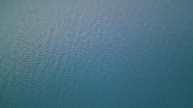 Aerial close-up view of sunlit sea surface with gentle ripples and deep green-blue tones. Natural ocean water texture with sunlight reflection and subtle wave patterns