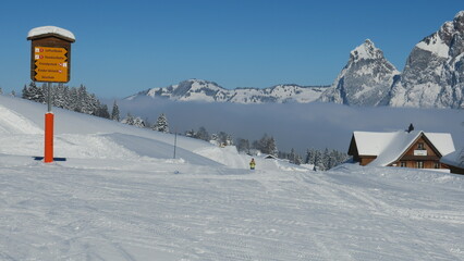 Fototapeta premium Scenic winter landscape: snow-covered mountains, ski slope, cozy cabin, and a trail sign.