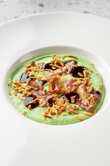 A gourmet dish featuring a vibrant green puree, crispy bacon, fried onions, and a rich dark sauce, elegantly plated on a white dish against a textured grey background.