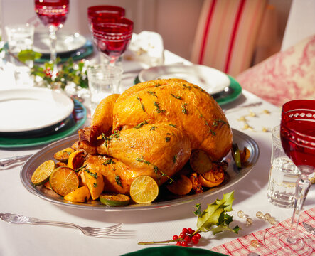 A beautifully set dinner table showcases a golden roast turkey, garnished with herbs and citrus on a platter. Surrounding dishes and decorations highlight the festive atmosphere.