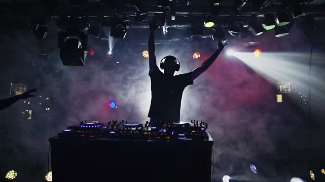 Silhouetted DJ in a smoky club, backlit by dramatic lighting. Captured from a low angle, this video evokes a vibrant nightlife atmosphere.