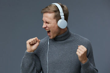 Energetic young man wearing white headphones and a gray turtleneck sweater enthusiastically...