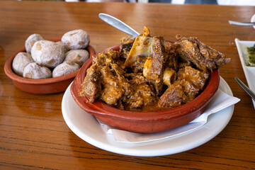 A bowl of goat meat with potatoes and a spoon on top. The goat meat is browned and the potatoes are...
