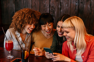 Four young women laughing and enjoying a night out at the bar, looking at smartphone
