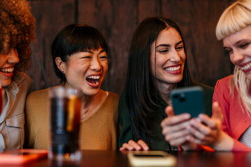 Cheerful female friends laughing and enjoying smartphone content in cafe