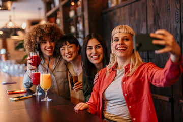 Four cheerful female friends taking selfie in bar while drinking cocktails