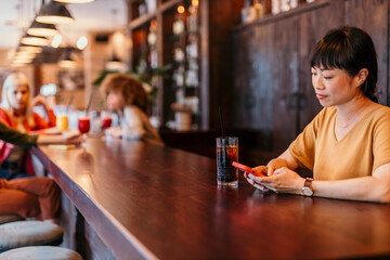 Woman using smartphone and drinking cocktail in bar counter with friends in background