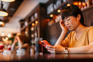 Stressed asian woman having headache reading bad news on smartphone in cafe