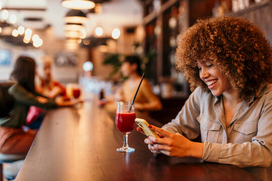 Cheerful young woman using smartphone and drinking juice at bar counter - Powered by Adobe
