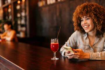 Cheerful young woman using smartphone and drinking juice at cafe bar counter