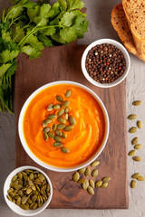 A vibrant bowl of creamy pumpkin soup garnished with pumpkin seeds, surrounded by multicolor peppercorns, fresh cilantro, rustic bread, and extra seeds for topping on a wooden board background