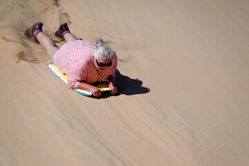 Older solo senior woman sandboarding down sand dune on boogie board, retirement fun excitement, adventure travel, holiday vacation activity, healthy vital age ageing © Cynthia