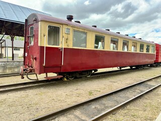 Fototapeta premium Old red and beige passenger train car at a rural station under cloudy skies, symbolizing travel history.