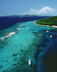 Sailboats anchored near tropical coral reef and forested coastline