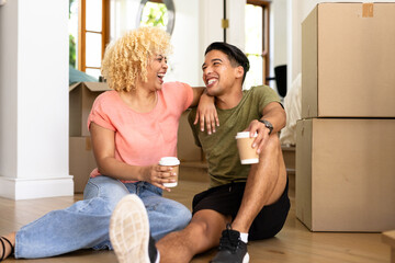 Young diverse couple sitting on floor with coffee, smiling and unpacking moving boxes