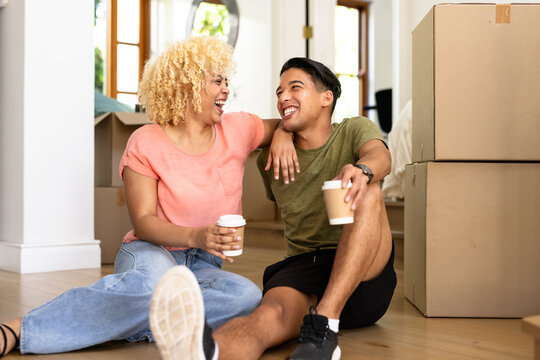 Young diverse couple sitting on floor with coffee, smiling and unpacking moving boxes - Powered by Adobe