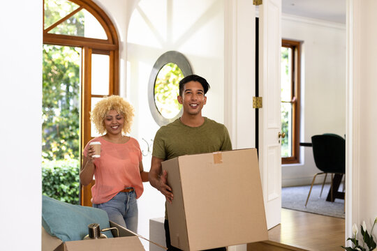 Young diverse couple carrying boxes and coffee, moving into new home, smiling happily