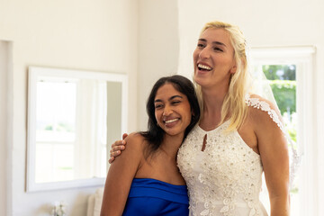 Bride and friend smiling joyfully in elegant wedding attire at home
