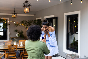 Naklejka premium Man proposing to surprised woman on patio, romantic moment under string lights