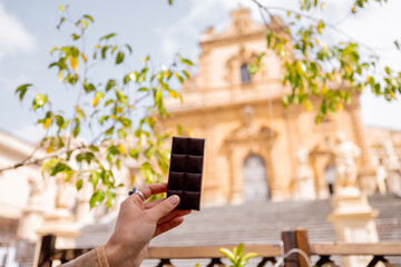 Close-up of a hand holding traditional Modica chocolate bar with blurred view of Saint Peter...