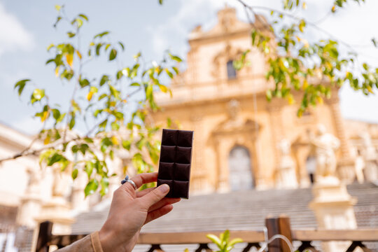 Close-up of a hand holding traditional Modica chocolate bar with blurred view of Saint Peter Cathedral in Modica, Sicily, a tribute to authentic taste and heritage