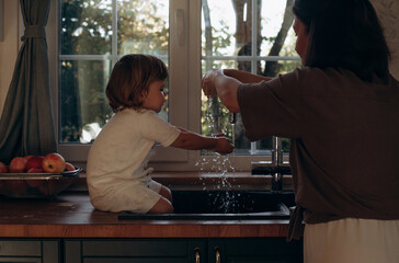 A child sits in the kitchen sink splashing water as a parent washes apples nearby. Sunlight streams through the window, creating a warm atmosphere of family bonding and play