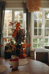 This interior setting features a stylish autumn-themed floral arrangement on a wooden dining table, capturing the warm ambiance of a sunlit afternoon. Windows showcase lush greenery outside