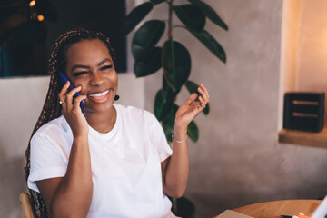 Close-up of cheerful young Black woman laughing during phone conversation, sitting in cozy workspace with relaxed attitude and tech gear in background