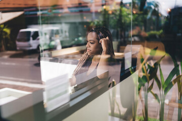 Pensive young African American woman with braids sitting by cafe window, looking outside in deep thought. Reflection in glass, natural light, casual clothes, moment of focus and contemplation