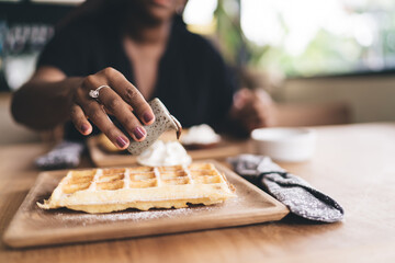 Close-up of African American woman’s hand pouring chocolate sauce over freshly made Belgian waffles with whipped cream, preparing her sweet brunch at a cozy café table