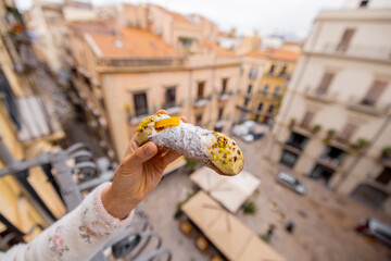 A hand holds a traditional Sicilian cannolo over a balcony with a view of old Palermo rooftops. This image blends iconic local cuisine with travel storytelling, ideal for food, tourism