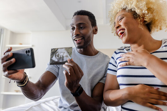 Excited couple holding ultrasound photo while video calling family at home