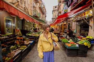 Obraz premium A woman in a yellow jacket strolls through a vibrant local market in Sicily, surrounded by fresh produce. The image conveys authentic travel, cultural immersion, and slow lifestyle 
