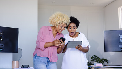 Two women excitedly looking at tablet and smartphone in modern office