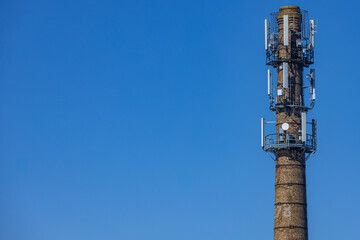 A repurposed brick smokestack outfitted with multiple modern cellular antennas and satellite dishes, set against a vibrant clear blue sky. Includes generous copy space.