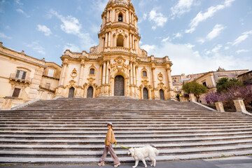 Woman walks her large white dog past the grand staircase of the Cathedral of San Giorgio in Modica, Sicily, under a bright blue sky